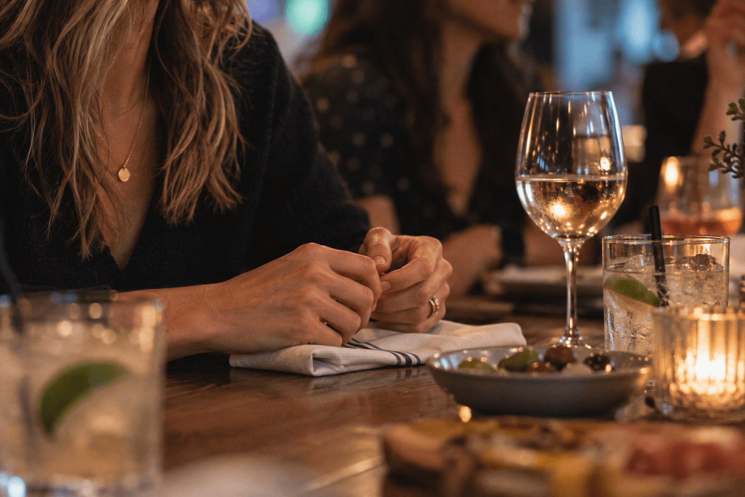 Close editorial photograph of a woman's hands fidgeting with a napkin at a restaurant table while alcoholic drinks are nearby.